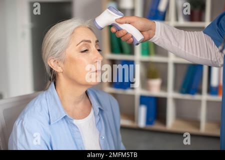 Der Arzt misst die Temperatur mit einem berührungslosen Thermometer bei einer alten Kaukasierin in der Klinik Stockfoto