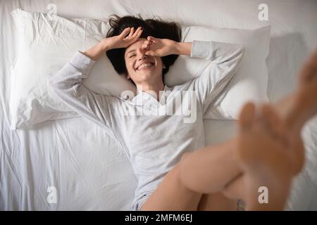 Lachende junge europäische Frau wacht auf, erhebt die Beine, hat allein Spaß, genießt guten Morgen, Urlaub Stockfoto