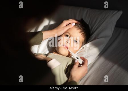 Der traurige kleine Junge liegt auf dem Bett, die mutter misst die Temperatur mit einem Thermometer und berührt die Stirn im Schlafzimmer Stockfoto