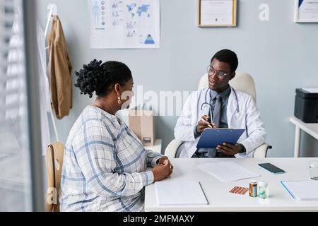 Porträt eines jungen schwarzen Arztes, der eine junge Frau in der Klinik konsultiert und auf Symptome hört Stockfoto