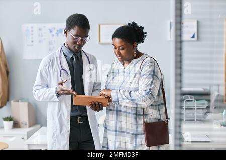 Taillenhochporträt einer jungen schwarzen Arztin, die eine Patientin aufsucht und in der Klinik ein digitales Tablett einsetzt Stockfoto