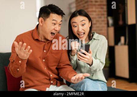 Porträt eines überfreulichen asiatischen Paares, das ein Smartphone benutzt und eine Siegergeste macht, die Fäuste schüttelt, zu Hause auf der Couch sitzt Stockfoto