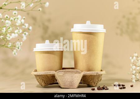 Kaffee zum Mitnehmen. Frühlingszusammensetzung mit zwei umweltfreundlichen Einwegbechern aus Pappe mit Kaffeebohnen auf beigefarbenem Hintergrund mit weißen Blumen. Co. Zum Mitnehmen Stockfoto