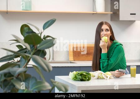 Eine attraktive Brünette hält einen grünen Apfel, während sie am Küchentisch steht. Stockfoto