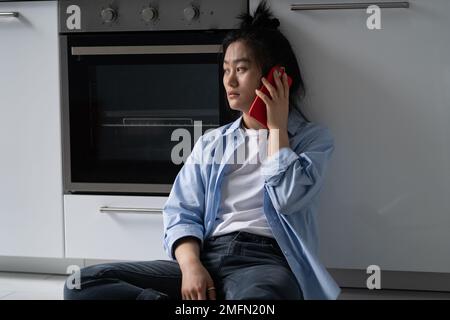 Traurige, entmutigte Asiatin, die versucht, ihren Freund anzurufen, sitzt auf dem Boden in der Küche und erlebt Stress Stockfoto