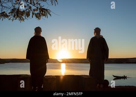 Zwei junge arabische Männer, die Djellaba und Turban tragen und den Sonnenuntergang vor einem See am klaren Himmel beobachten Stockfoto