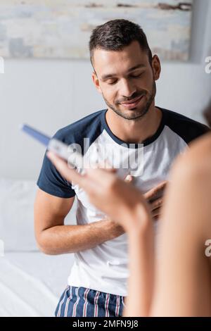 Lächelnder Mann im Pyjama, der neben der Freundin steht, mit Schwangerschaftstest im Schlafzimmer, Stockbild Stockfoto