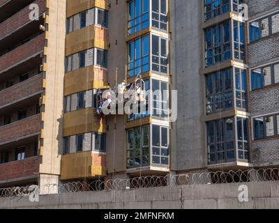 Männlicher Baumeister in einer Hängebrücke arbeitet an der Fassade eines neu gebauten Hochhauses. Neuer Bezirk im Bau in Kiew. Stockfoto