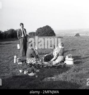 1950er, historisch, ein Familienpicknick, auf einem Hügel, ein Vater in Anzug und Krawatte, mit seiner Familie, die auf einem Teppich auf dem Gras sitzt, England, Großbritannien. Teekanne, Korb, Flasche, alles auf dem Bild. Stockfoto