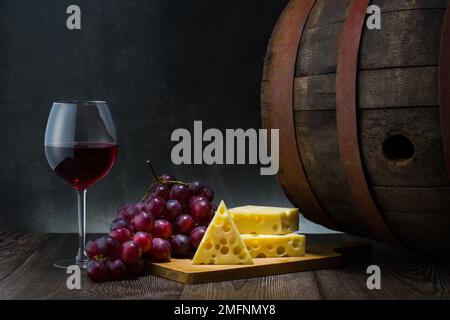 Rotwein bei schwachem Licht mit Käse und Trauben, Weinfass und Glas, Stille Leben Stockfoto