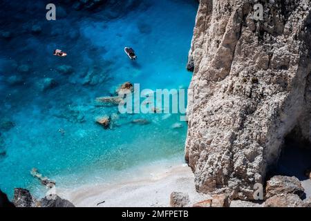 Touristen schnorcheln im kristallklaren Wasser und ankern Boote neben einer zerklüfteten Felswand am Strand von Myzithres, Zakynthos, Griechenland Stockfoto