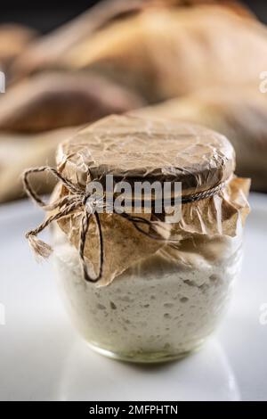 Ein Glas mit hausgemachtem Roggen-Sauerteig und frischem Brot im Hintergrund. Stockfoto
