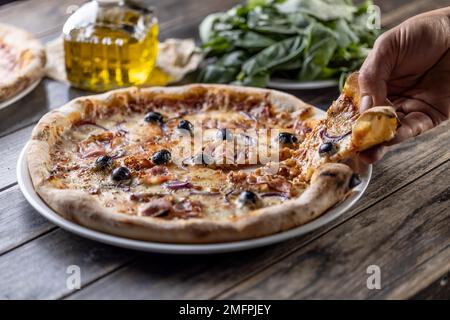 Die Hand eines Mannes hält das erste Stück frisch gebackener Pizza, frisches Basilikum und eine Karaffe Olivenöl im Hintergrund. Stockfoto