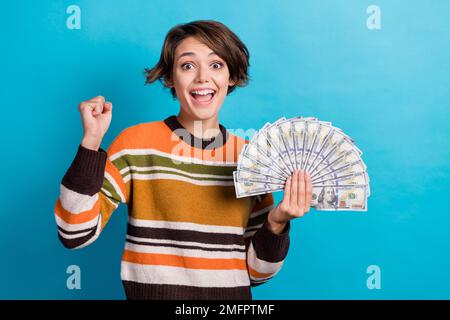 Foto einer lustigen Frau mit gestreiftem Pullover mit Bargeld, die aufsteigende Faust, isolierter blauer Hintergrund Stockfoto