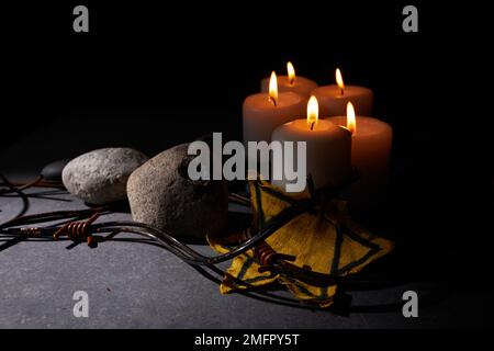 Holocaust-Gedächtnisfeiertag. Stacheldraht, Steine, gelber Stern und brennende Kerze auf schwarzem Hintergrund Stockfoto