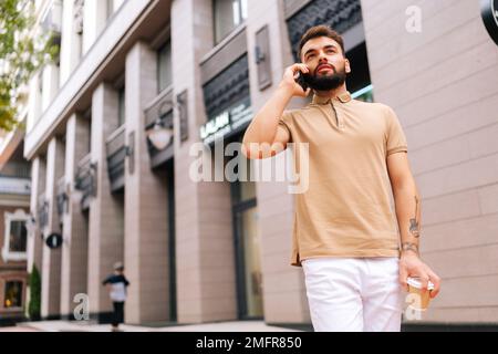 Blick auf einen gutaussehenden jungen Mann mit tätowierter Hand, der auf der Straße spaziert, mit dem Smartphone spricht und Kaffee zum Mitnehmen trinkt. Stockfoto