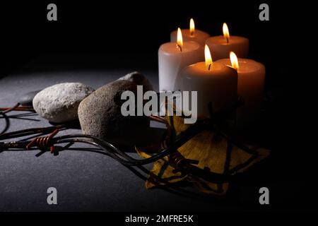 Holocaust-Gedächtnisfeiertag. Stacheldraht, Steine, gelber Stern und brennende Kerze auf schwarzem Hintergrund Stockfoto