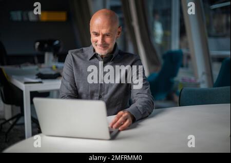 Ein Geschäftsmann mittleren Alters, der ein Notebook benutzt, während er am Schreibtisch sitzt und in einem modernen Büro arbeitet. Stockfoto
