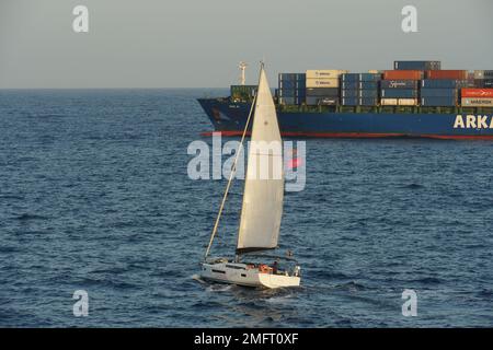 Weißes Segelboot, das am Anker vor dem Hafen von Valencia im Mittelmeer um das blaue Containerschiff vorbeifährt. Stockfoto