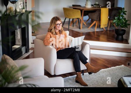 Eine lächelnde Geschäftsfrau mittleren Alters, die ihr Smartphone und Kopfhörer benutzt, während sie zu Hause am Sessel sitzt und ein Videogespräch führt. Stockfoto
