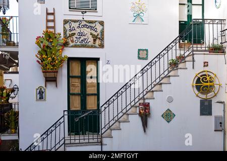 Weiße Fassade eines Hauses mit grünen Türen, Keramikfliesen und Blumen in Töpfen in einer der Gassen des Touristenziels Taormina. Stockfoto