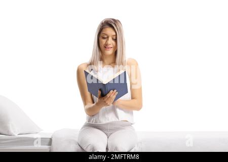 Frau im Pyjama, die auf einem Bett sitzt und ein Buch liest, isoliert auf weißem Hintergrund Stockfoto