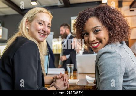 Junge schwarze südamerikanerin, die zusammen mit ihrem blonden Kaukasier in einem modernen Büro vor die Kamera schaut - Pause von der Arbeit und Zusammenarbeit Stockfoto