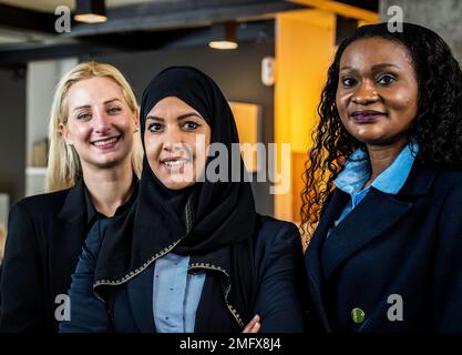 Lächelnde Geschäftsfrauen mit unterschiedlichen ethnischen Zugehörigkeiten und Kulturen, die in einem modernen Büro vor die Kamera schauen - drei elegante Freundinnen, eine davon trägt sie Stockfoto