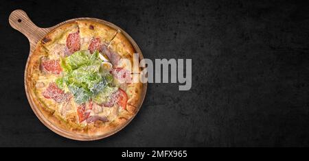 Pizza mit Schinken, Hühnchen, Tomaten, Salat und Käse, auf einem Holzbrett, auf grauem Beton Stockfoto