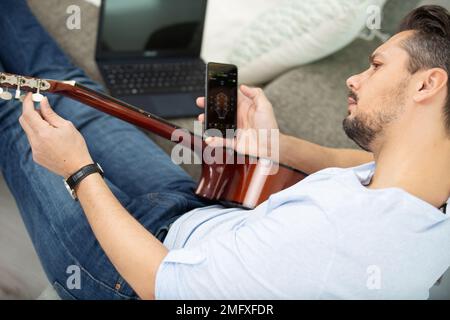 Ein junger Mann, der beim Gitarrenspielen ein Handy benutzt Stockfoto