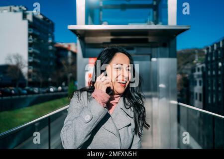 Lächelnde und fröhliche, brünette Frau, die im Business Center mit dem Handy telefoniert Stockfoto