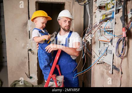 Kinderelektriker, der in der Leiter steht und das Zangenwerkzeug hält, während er die Schalttafel mit dem Vater zu Hause installiert. Mann und Kind bauen elektrische Leitungen in einer Wohnung, die gerade renoviert wird. Stockfoto