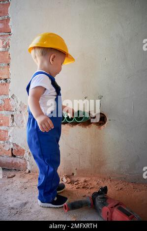 Ein kleiner Elektriker, der Kabelkanäle und Steckdosen in der Wand installiert. Kind im Sicherheits- und Arbeitsanzug Bauhelm Montage der elektrischen Verkabelung in einer Wohnung, die gerade renoviert wird. Stockfoto