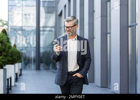 Erfolgreicher, grauhaariger Geschäftsmann geht vor das Bürogebäude, ein reifer Chef mit Brille benutzt Telefon, lächelt und schreibt Nachrichten, liest Online-Nachrichten und durchsucht Internetseiten. Stockfoto