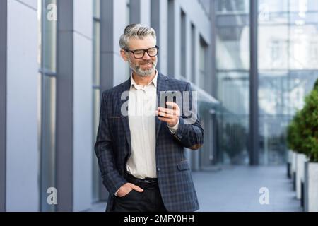 Erfolgreicher, grauhaariger Geschäftsmann geht vor das Bürogebäude, ein reifer Chef mit Brille benutzt Telefon, lächelt und schreibt Nachrichten, liest Online-Nachrichten und durchsucht Internetseiten. Stockfoto