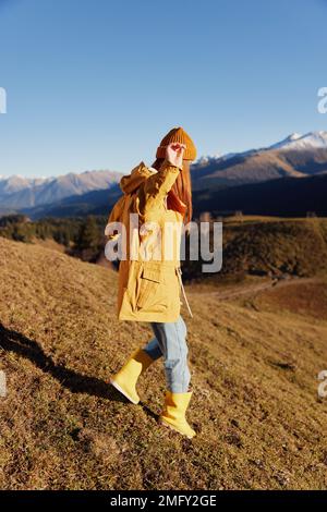 Die Frau geht auf einem Hügel in voller Länge spazieren und sieht die Berge in einem gelben Regenmantel und Jeans im Herbst auf einer schönen Reise bei Sonnenuntergang wandern Stockfoto