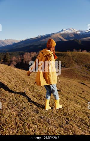 Mit dem Rücken einer Frau auf dem Hügel joggen und die Berge in einem gelben Regenmantel und Jeans im Herbst betrachten Happy Sunset Trip auf einem Stockfoto