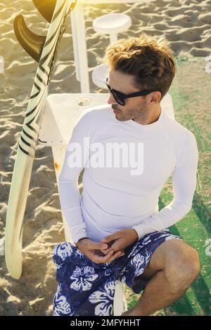 Ein junger gutaussehender, lächelnder Mann sitzt am Strand neben seinem Surfbrett und trägt eine dunkle Sonnenbrille und ein weißes, leeres, langärmeliges Lycra-Hemd. Vertikales Modell- Stockfoto