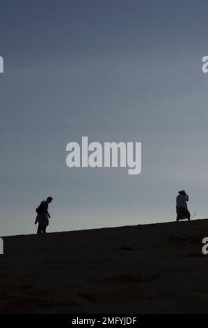 Hintergrundbeleuchtete Silhouetten von zwei Personen auf einer Sanddüne mit Sonnenaufgang hinter ihnen in Vietnam und einer Frau in einem traditionellen vietnamesischen Hut aus Bambus Stockfoto