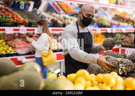 Latino-amerikanischer Arbeiter in Maske im Supermarkt mit Ananas während des Coronavirus-Pneumonieausbruchs Stockfoto