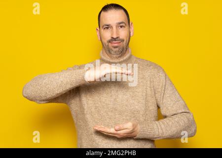 Ein Mann, der ein Objekt zwischen seinen Händen hält, ein Objekt an einer gelben Wand zeigt, anbietet oder anpreist. Leer. Leerzeichen. Konzept. Objekt. Unten Stockfoto