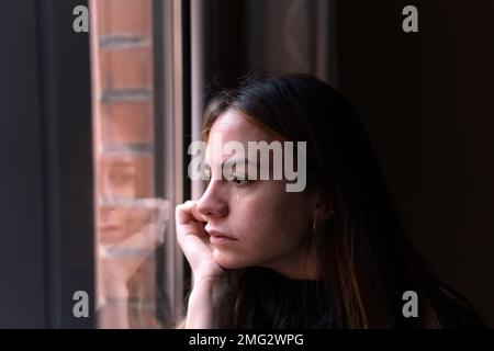 Seitenansicht einer einsamen Frau mit dunklen Haaren, die aus dem Fenster blickte und beim Weinen ihr Gesicht berührte Stockfoto