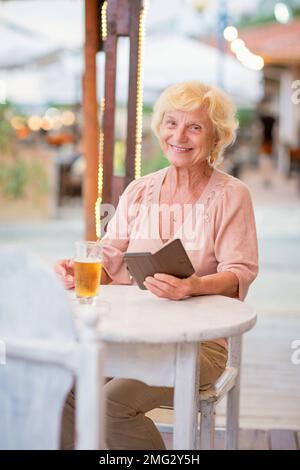 Reife Frau, die in einem Sommercafé an einem Tisch sitzt und Bier trinkt Stockfoto