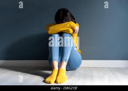 Der ganze Körper einer unbekannten, unglücklichen jungen Dame mit dunklen Haaren in legerer Kleidung umschließt das Knie und bedeckt das Gesicht, während sie auf dem Boden neben der Wand saß Stockfoto