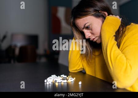 Seitenansicht einer melancholischen, schweren Frau mit langem Haar im gelben Pullover, die am Tisch sitzt und zuhause einen Haufen Antidepressiva-Pillen betrachtet Stockfoto