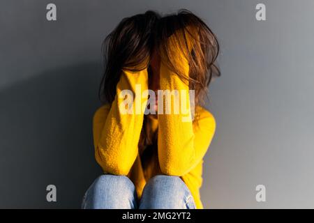 Verärgerte junge deprimierende Frau in Jeans und Pullover, die langes braunes Haar berührte, während sie auf dem Boden saß und sich an eine graue Wand lehnte Stockfoto