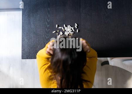 Draufsicht einer nicht erkennbaren deprimierenden Frau mit langem dunklem Haar im gelben Pullover, die den Kopf berührt, während sie mit einem Haufen Pillen am Tisch sitzt Stockfoto