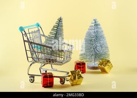 Kleiner Weihnachtsbaum in einem kleinen Einkaufswagen. Ein weiterer Weihnachtsbaum mit bunten Geschenken ist im Hintergrund. Weihnachtseinkaufskonzept. Speicherplatz kopieren Stockfoto