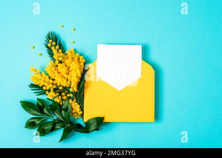 Umschlag mit leerer Karte, mimosagelbe Blumen auf blauem Hintergrund. Frühjahrsferien-Konzept. Draufsicht, flaches Lay, Modell. Stockfoto