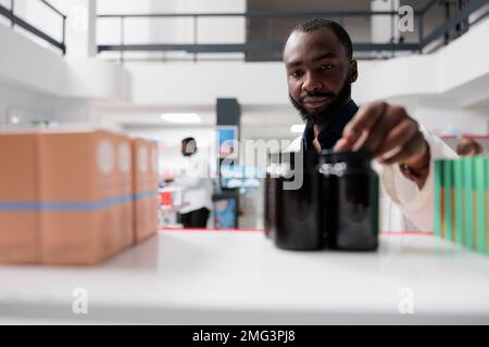 afroamerikanischer Apotheker nimmt Arzneimittelflaschen aus dem Apothekenregal, selektiver Fokus. Apotheke, die Pillen-Pakete aus der Nähe bringt, Supplemente verkauft, Vorderansicht Stockfoto